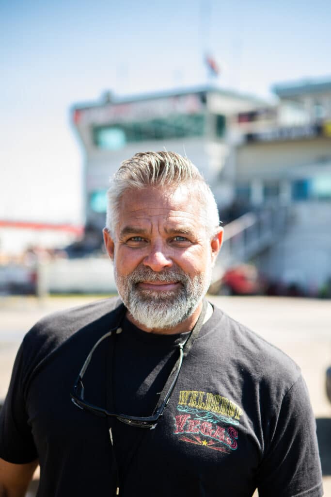 Close-up of a smiling older man with gray beard wearing a black T-shirt and sunglasses, outdoors on a sunny day.