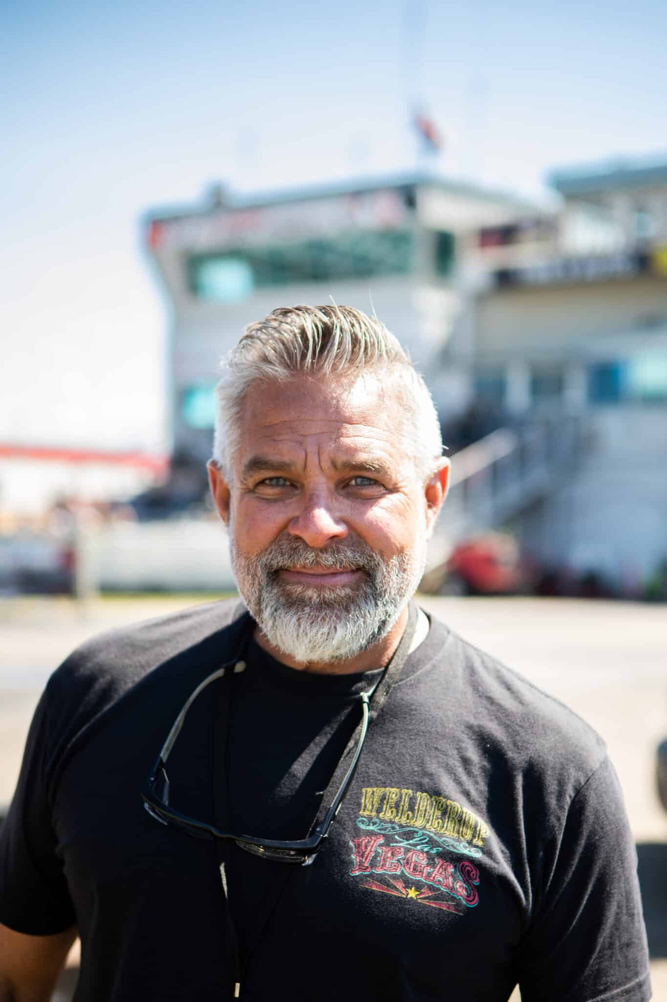 Close-up of a smiling older man with gray beard wearing a black T-shirt and sunglasses, outdoors on a sunny day.