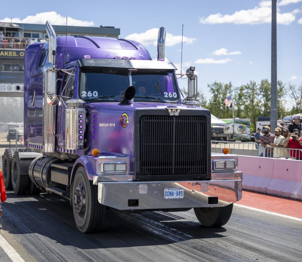 A purple semi-truck at a drag race track with spectators in the background and a man in a red shirt in the foreground.