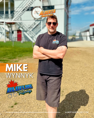 Mike Wynnyk with arms crossed, wearing a black T-shirt and sunglasses, posing on a gravel racing facility with 'MIKE WYNNYK' text and BlueLine Racing logo nearby.